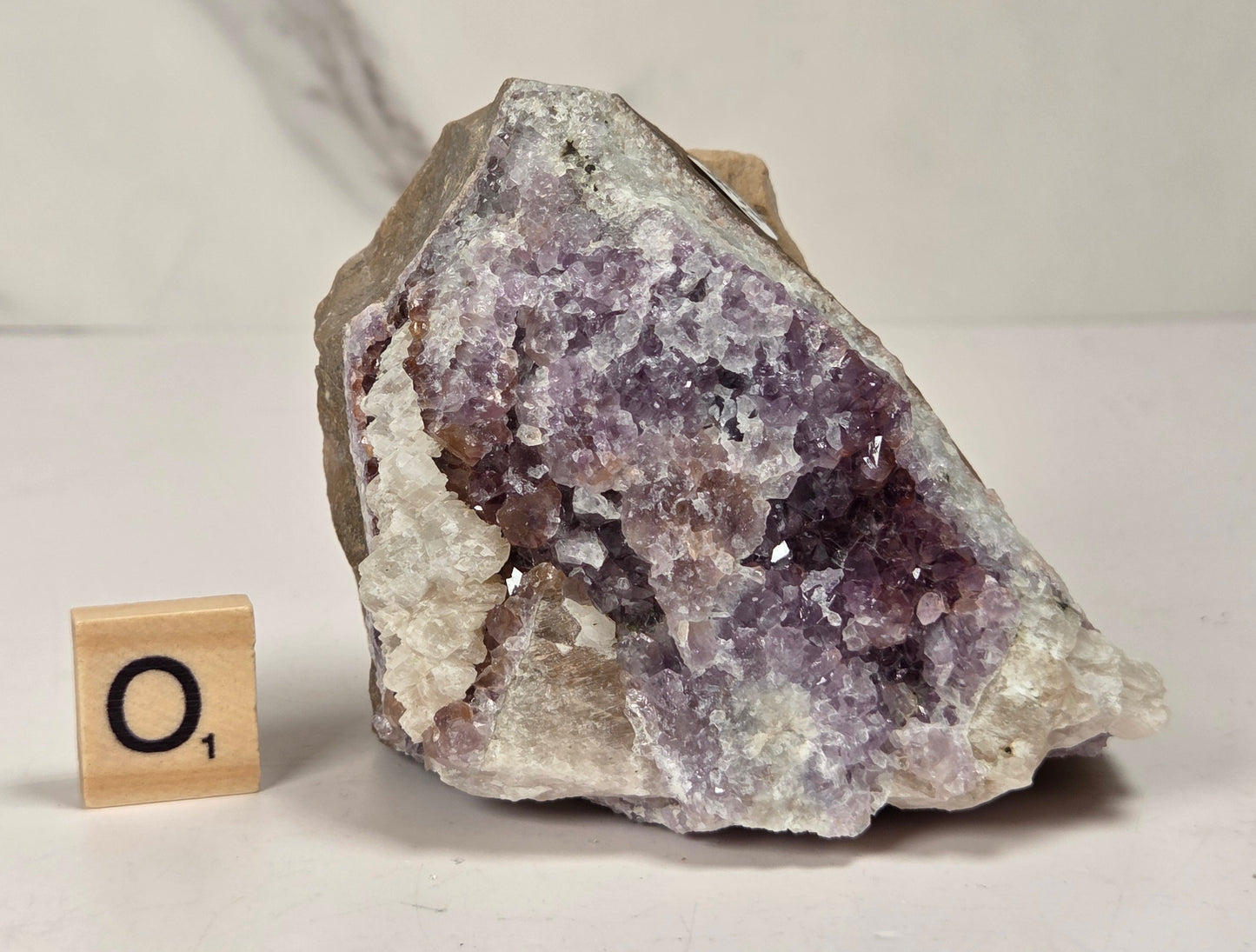 A raw amethyst geode specimen with purple crystal clusters and white matrix, displayed on a light surface next to a small wooden 'O1' tile.