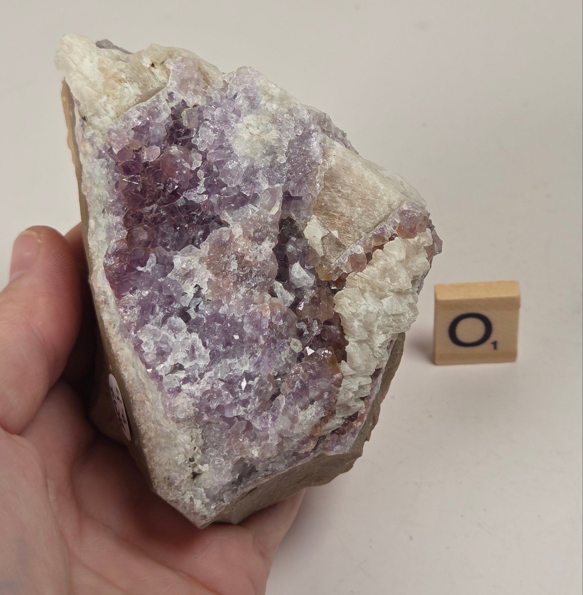 A hand holds a rough amethyst crystal cluster with purple and white crystalline formations. A small wooden tile with the letter 'O' sits on the beige background next to the specimen.