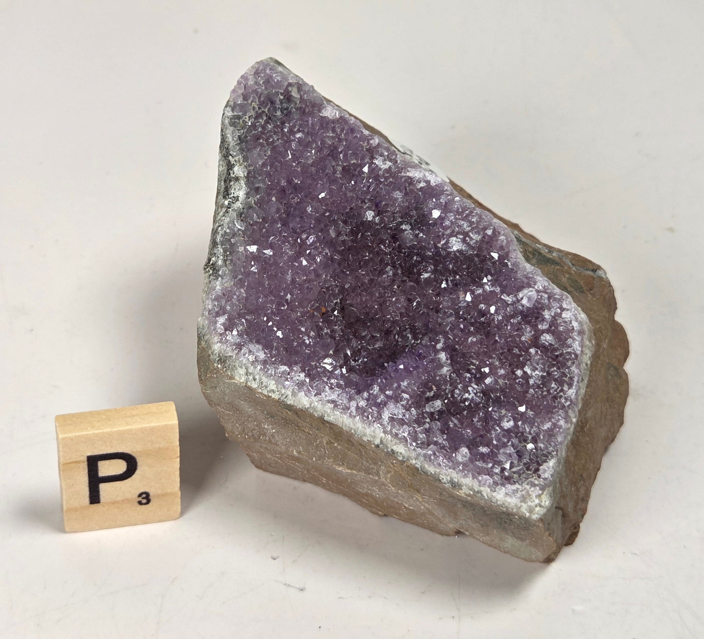 A raw amethyst geode specimen with a rough brown matrix, shown next to a small wooden Scrabble tile marked P3 on a light gray background.