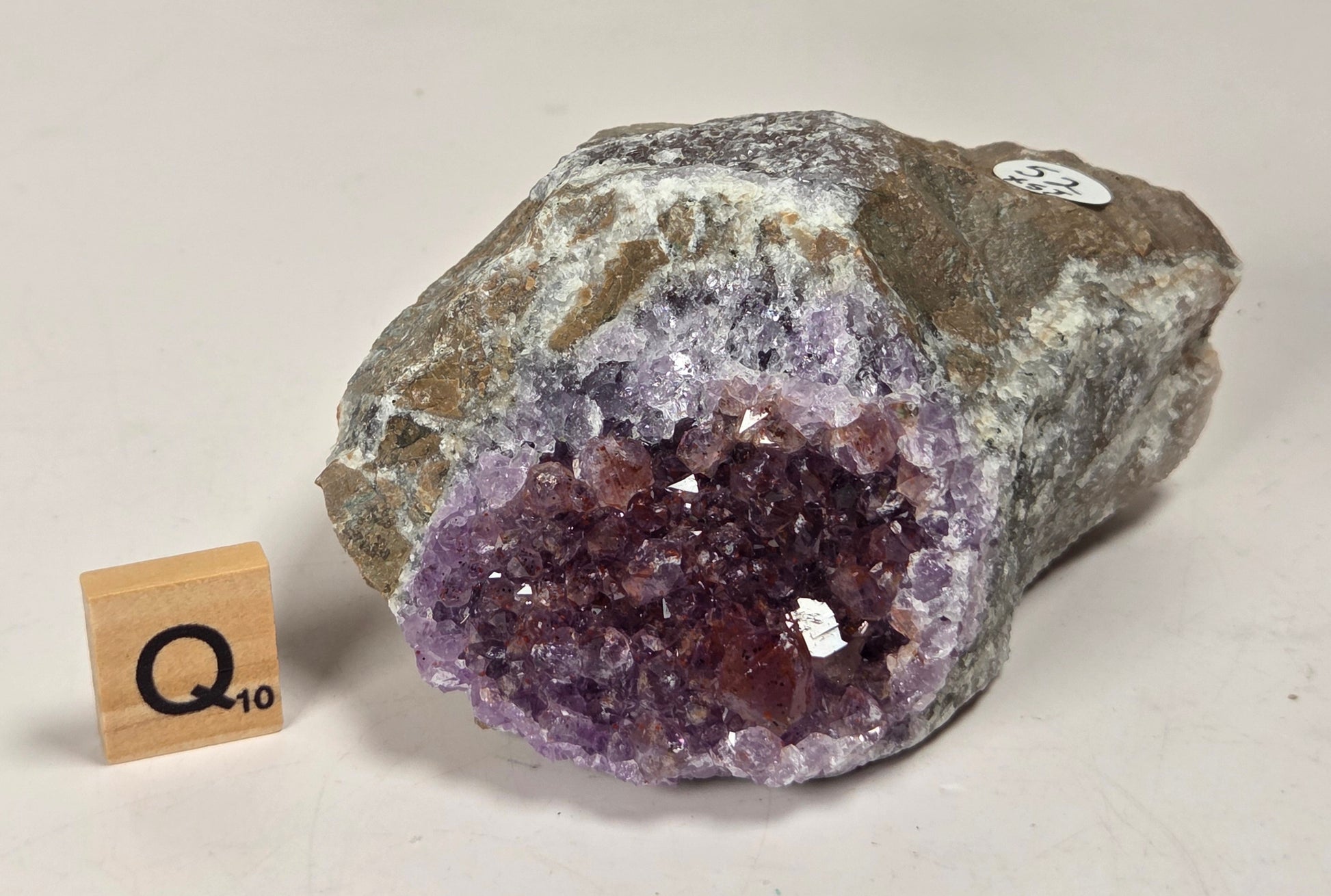 A raw amethyst geode specimen with a purple crystal cavity and rough gray-brown outer matrix, photographed on a plain light background. A small wooden tile with 'Q 10' is placed beside it for scale.