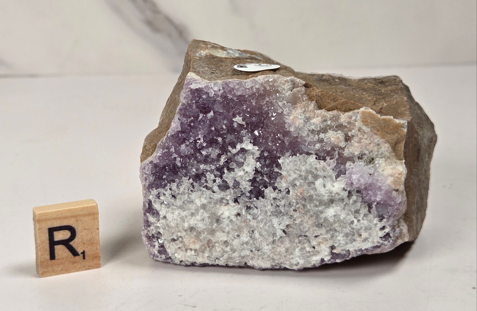 A raw amethyst crystal cluster with purple and white crystalline formations and a brown rough outer matrix, displayed on a light surface next to a small wooden letter tile.