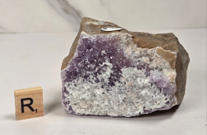 A raw amethyst crystal cluster with purple and white crystalline formations and a brown rough outer matrix, displayed on a light surface next to a small wooden letter tile.
