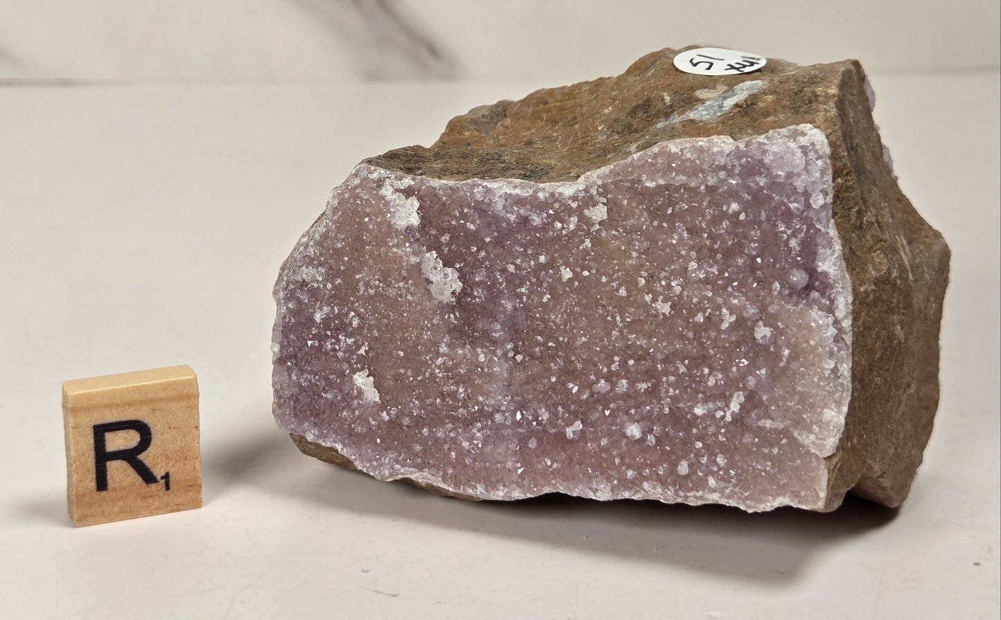 A raw amethyst geode specimen with a rough brown matrix and pale lavender-purple crystalline interior, shown on a light surface with a small wooden letter block displaying 'R1' for size reference.