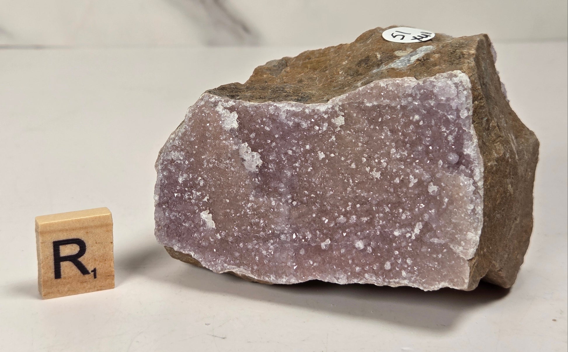 A raw amethyst geode specimen with a rough brown matrix and pale lavender-purple crystalline interior, shown on a light surface with a small wooden letter block displaying 'R1' for size reference.