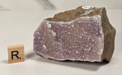 A raw amethyst geode specimen with a rough brown matrix and pale lavender-purple crystalline interior, shown on a light surface with a small wooden letter block displaying 'R1' for size reference.