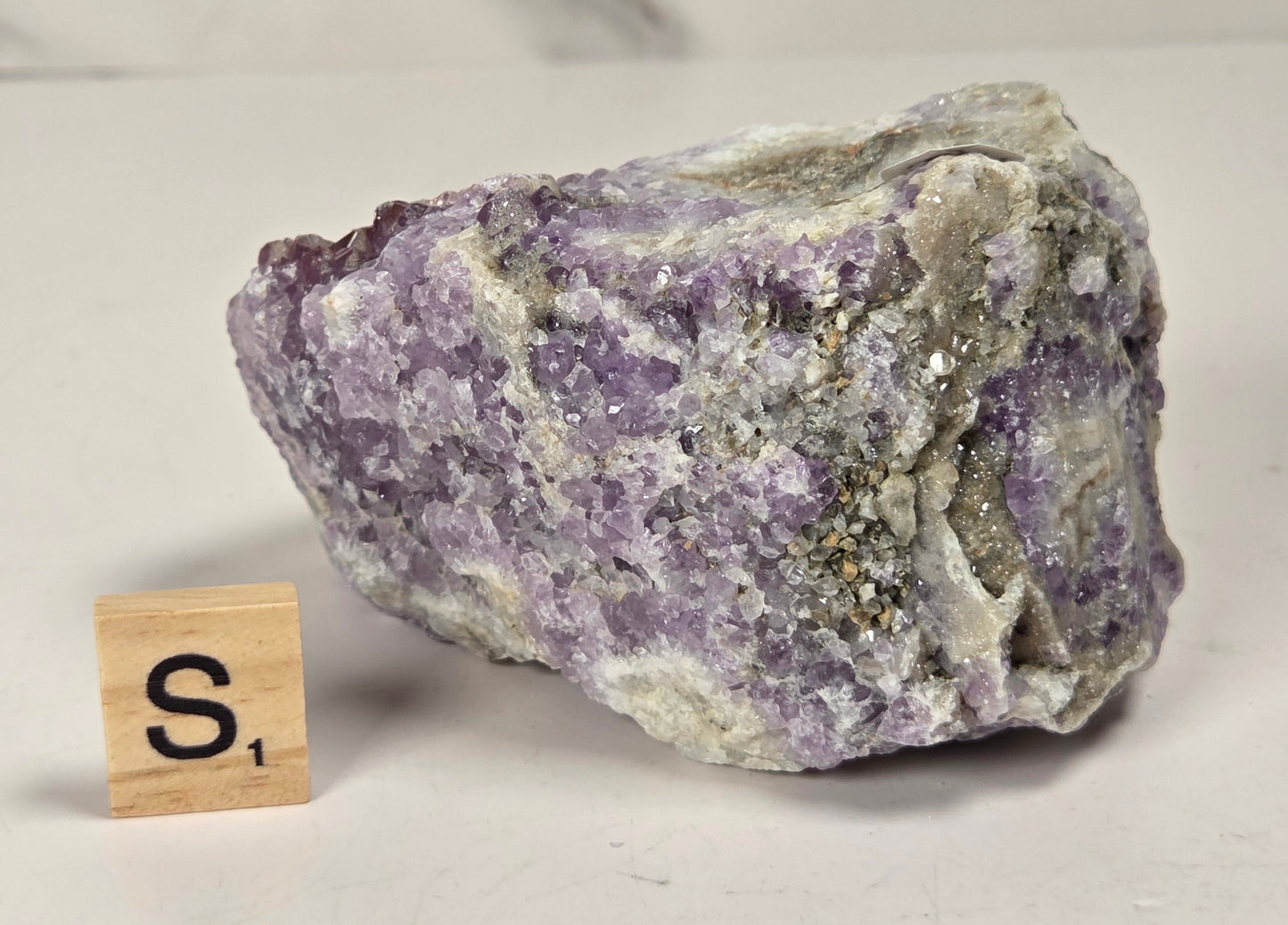 A rough purple amethyst crystal cluster with white and gray matrix, shown on a light surface next to a small wooden Scrabble tile with the letter S.