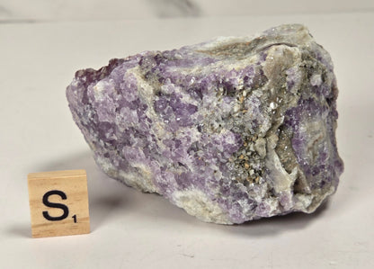 A rough purple amethyst crystal cluster with white and gray matrix, shown on a light surface next to a small wooden Scrabble tile with the letter S.