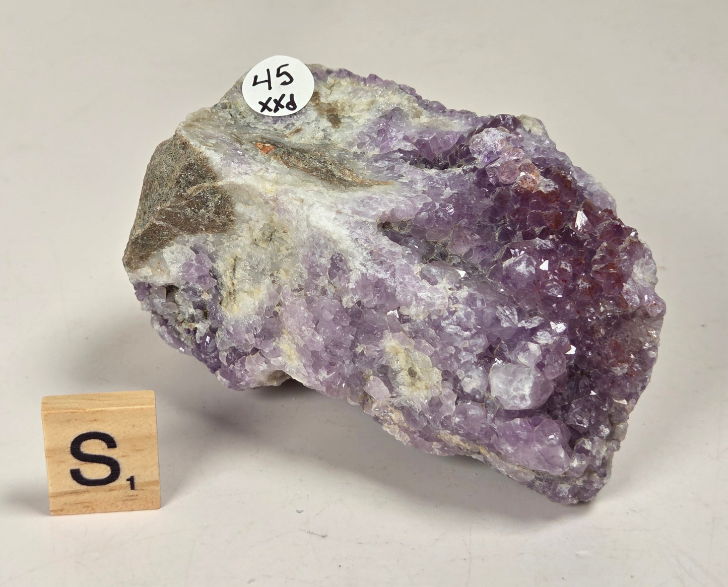 A raw amethyst crystal cluster with purple, lavender, and white crystalline formations and some gray-brown matrix, photographed on a light neutral surface. A small white sticker reading '45 XX4' is attached near the top, and a small wooden tile with the letter 'S' sits beside it.