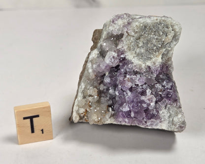 A raw amethyst crystal cluster with purple and white quartz points on a light gray surface, shown next to a small wooden Scrabble tile with the letter T.