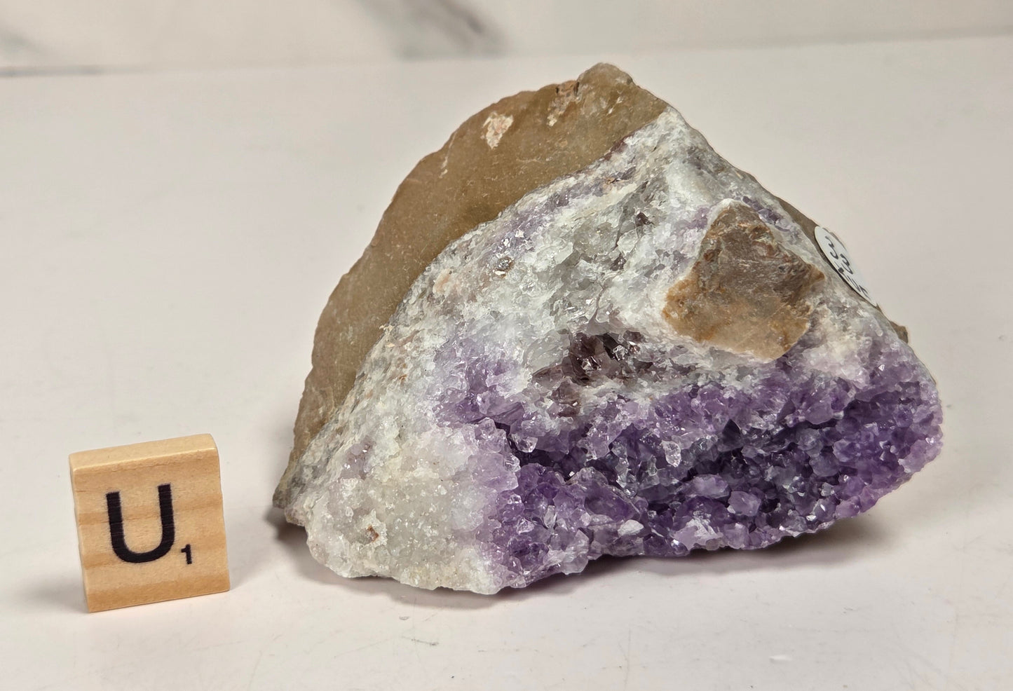 A raw amethyst crystal cluster with rough brown matrix, shown next to a small wooden letter tile displaying the letter 'U' on a light gray surface.