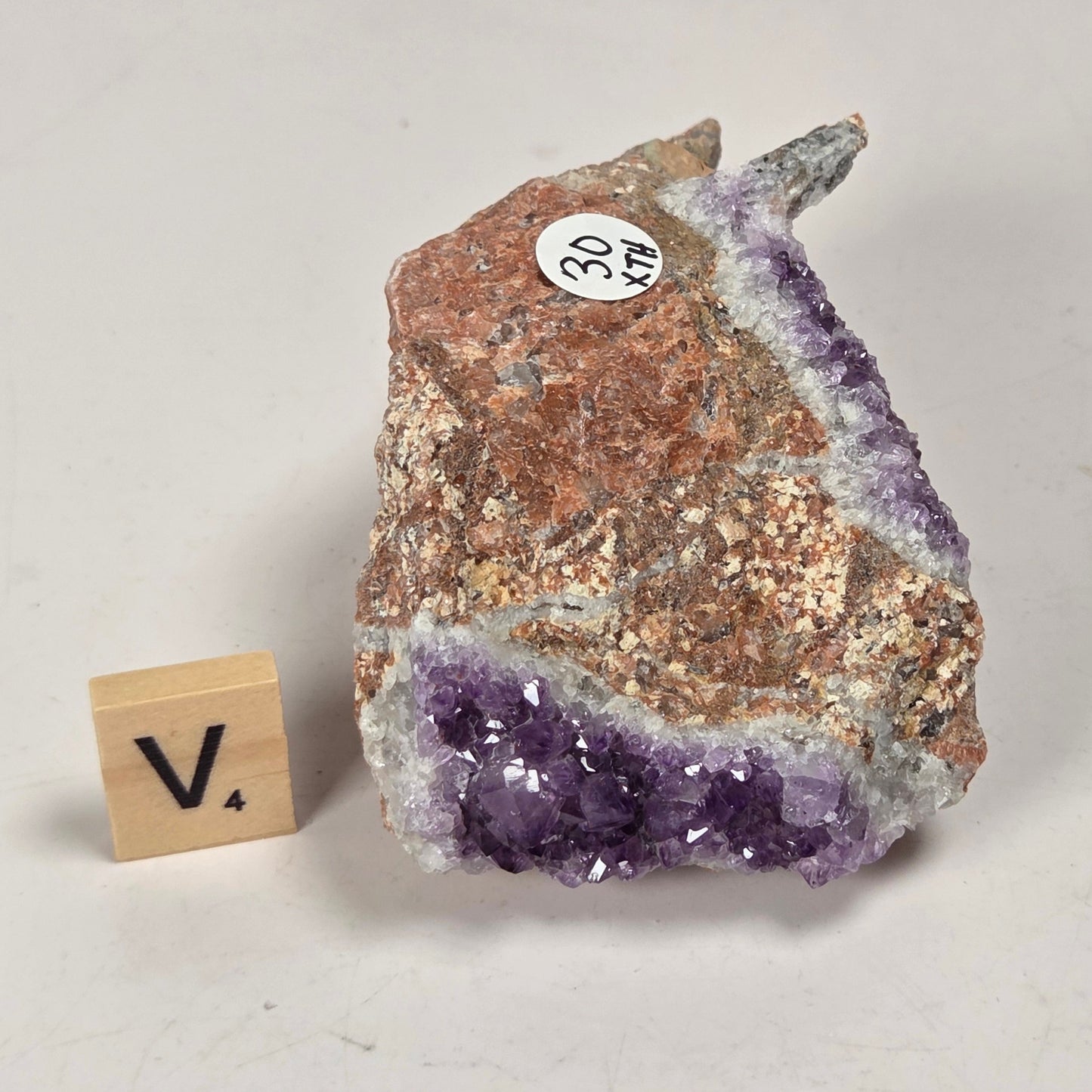 A raw amethyst geode specimen with rough brown and white crystalline matrix, shown on a light gray background next to a small wooden block with the letter V.