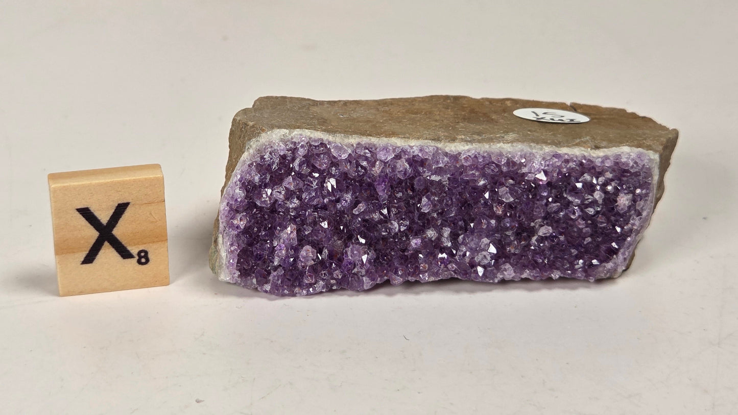 A raw amethyst crystal cluster specimen with a rough brown matrix, shown next to a wooden Scrabble tile displaying the letter 'X' and value 8, on a plain off-white background.