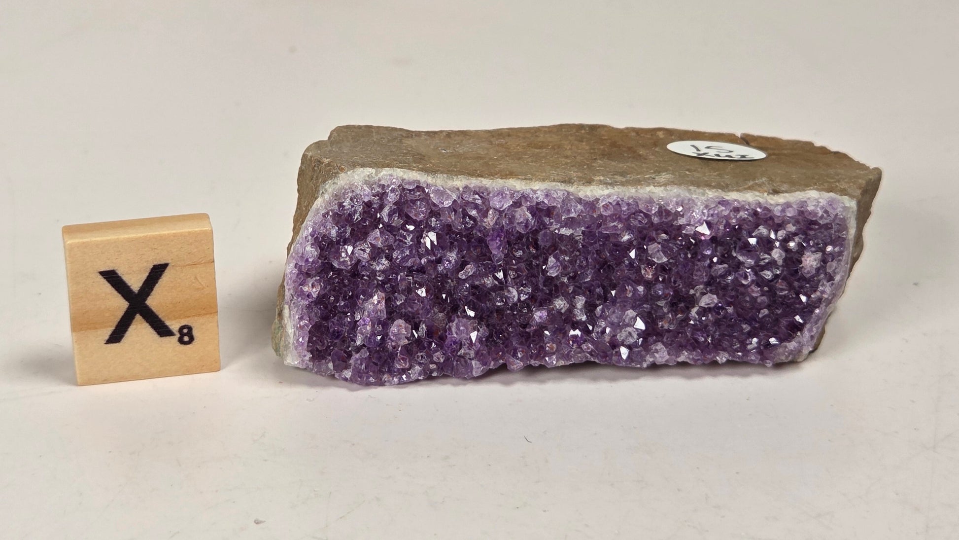 A raw amethyst crystal cluster specimen with a rough brown matrix, shown next to a wooden Scrabble tile displaying the letter 'X' and value 8, on a plain off-white background.