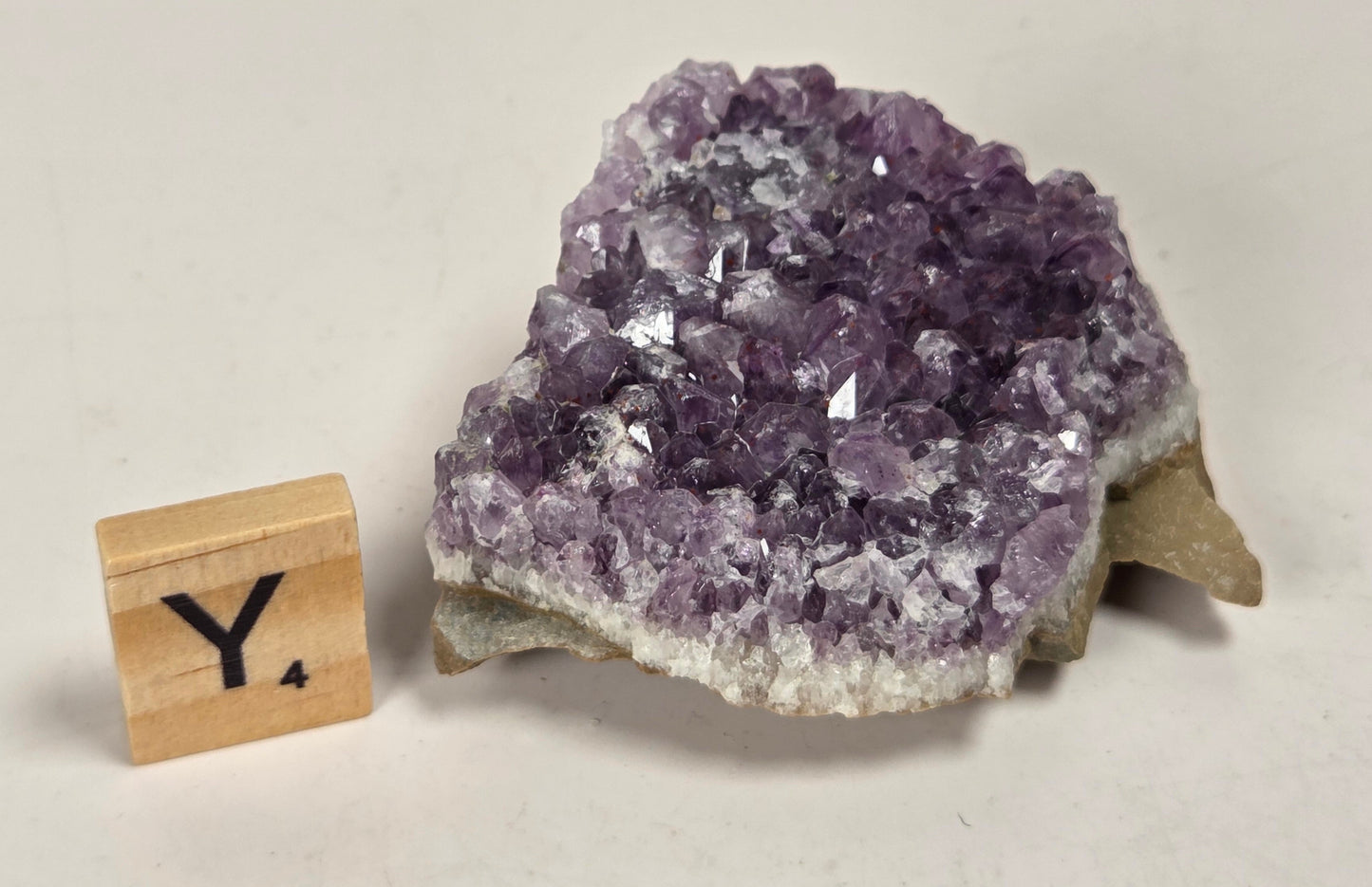 A natural amethyst crystal cluster with a rough matrix, shown next to a small wooden block letter 'Y' on a light beige background.