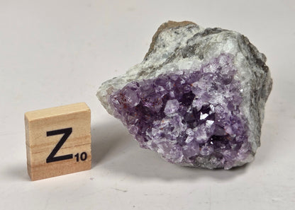A raw amethyst crystal cluster with purple crystalline points and a gray-brown rough matrix, shown next to a wooden Scrabble tile marked Z 10 on a light neutral background.