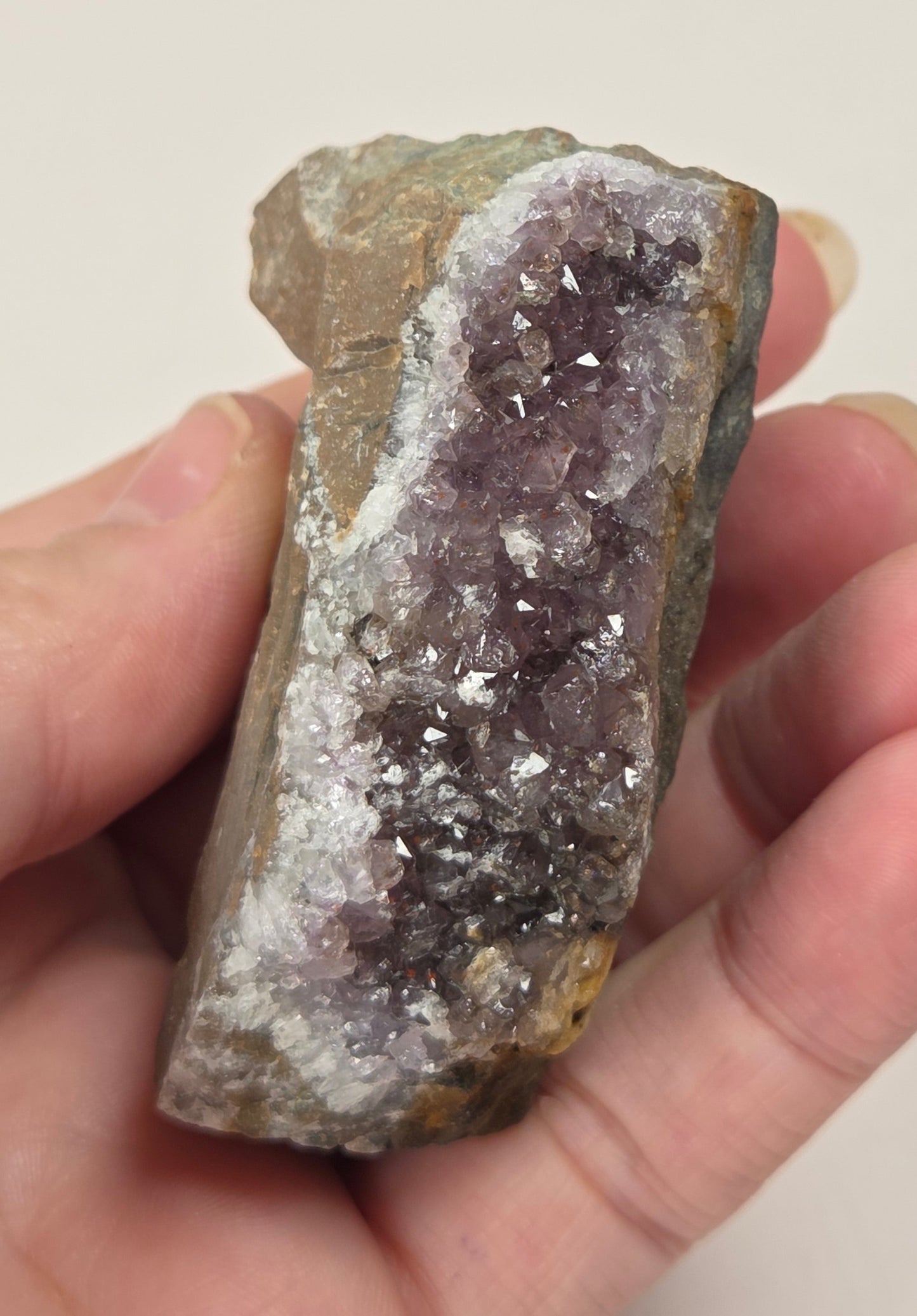 A hand holding a raw amethyst geode slice with purple crystal clusters and a brown, rough matrix.