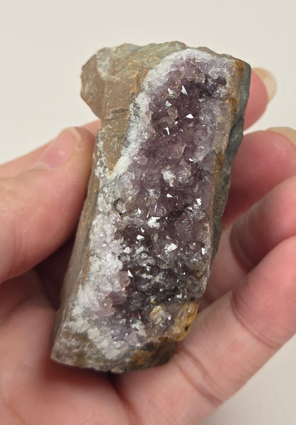 A hand holding a raw amethyst geode slice with purple crystal clusters and a brown, rough matrix.