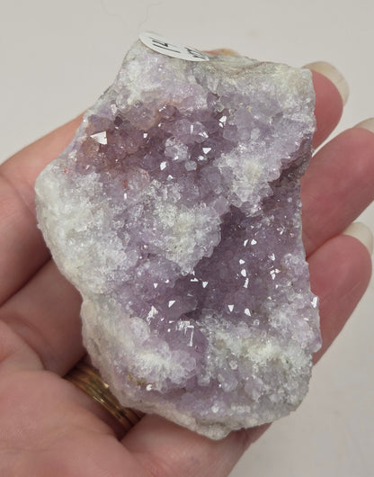 A hand holding a raw amethyst crystal cluster with light purple and white crystalline formations against a plain light background.