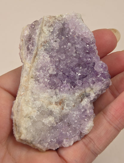 A hand holding a raw amethyst crystal cluster with purple and white crystalline formations against a plain beige background.