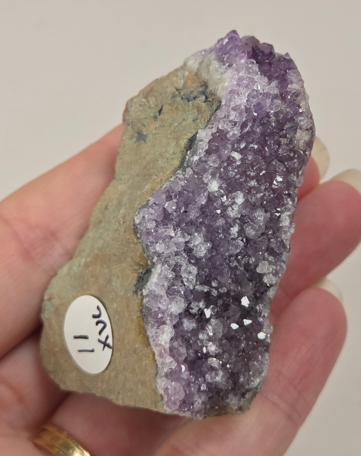 A hand holding a raw amethyst crystal cluster with rough matrix, showing a purple crystalline section and a tan-gray natural stone section with a small white label.