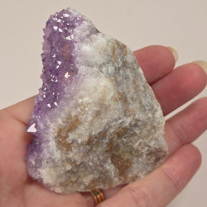 A raw amethyst crystal cluster held in a person's hand. The crystal features a prominent purple crystal cavity and outer crystal points on one side, transitioning into a rough, textured white and light brown matrix on the other.