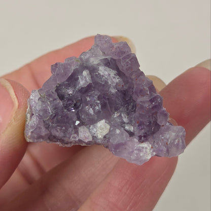 A hand holding a rough purple amethyst crystal cluster against a plain light background.