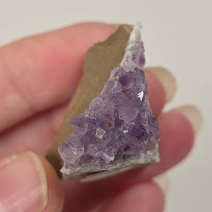 A hand holding a triangular crystal specimen with a cluster of light purple amethyst points on a tan and gray matrix, photographed against a plain light background.