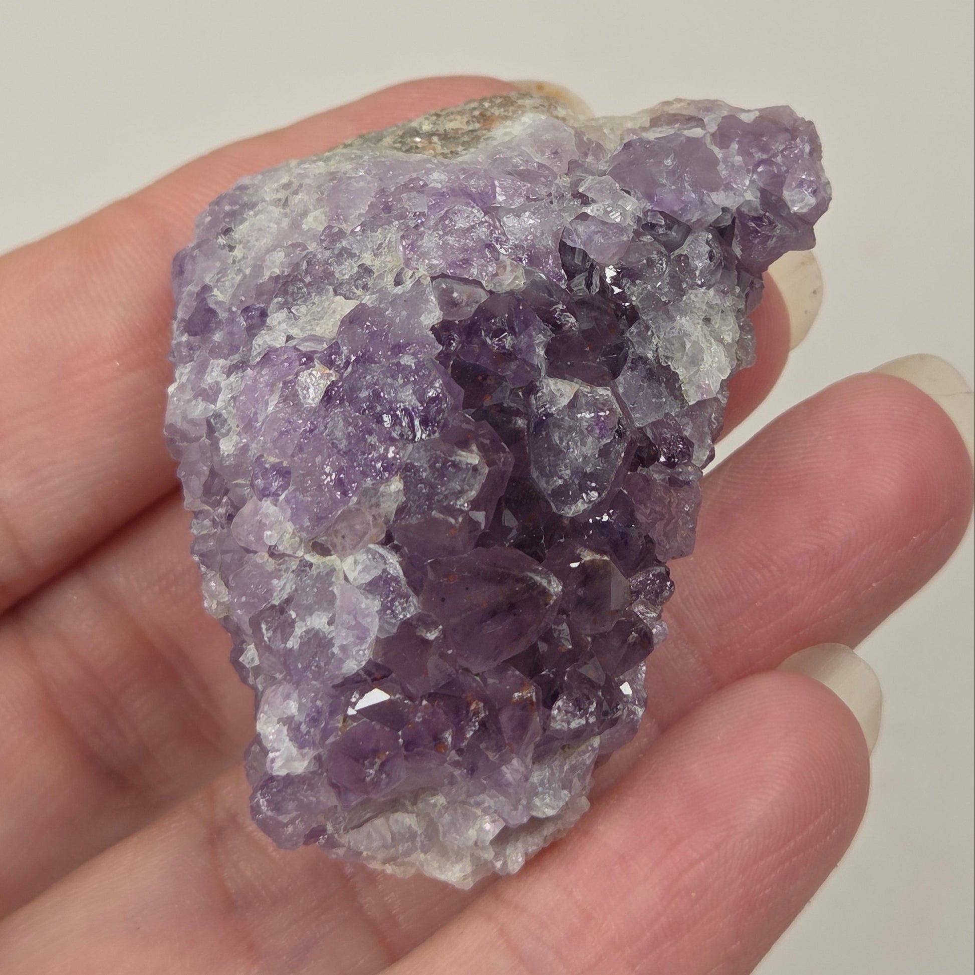 A hand holding a rough purple amethyst crystal cluster with visible natural facets and a light-colored matrix background, against a plain light gray backdrop.