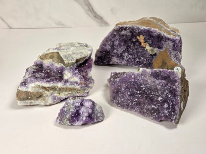 Four raw amethyst geode specimens with rough brown matrix and white crystalline surfaces, arranged on a white surface against a light gray marble-patterned background.