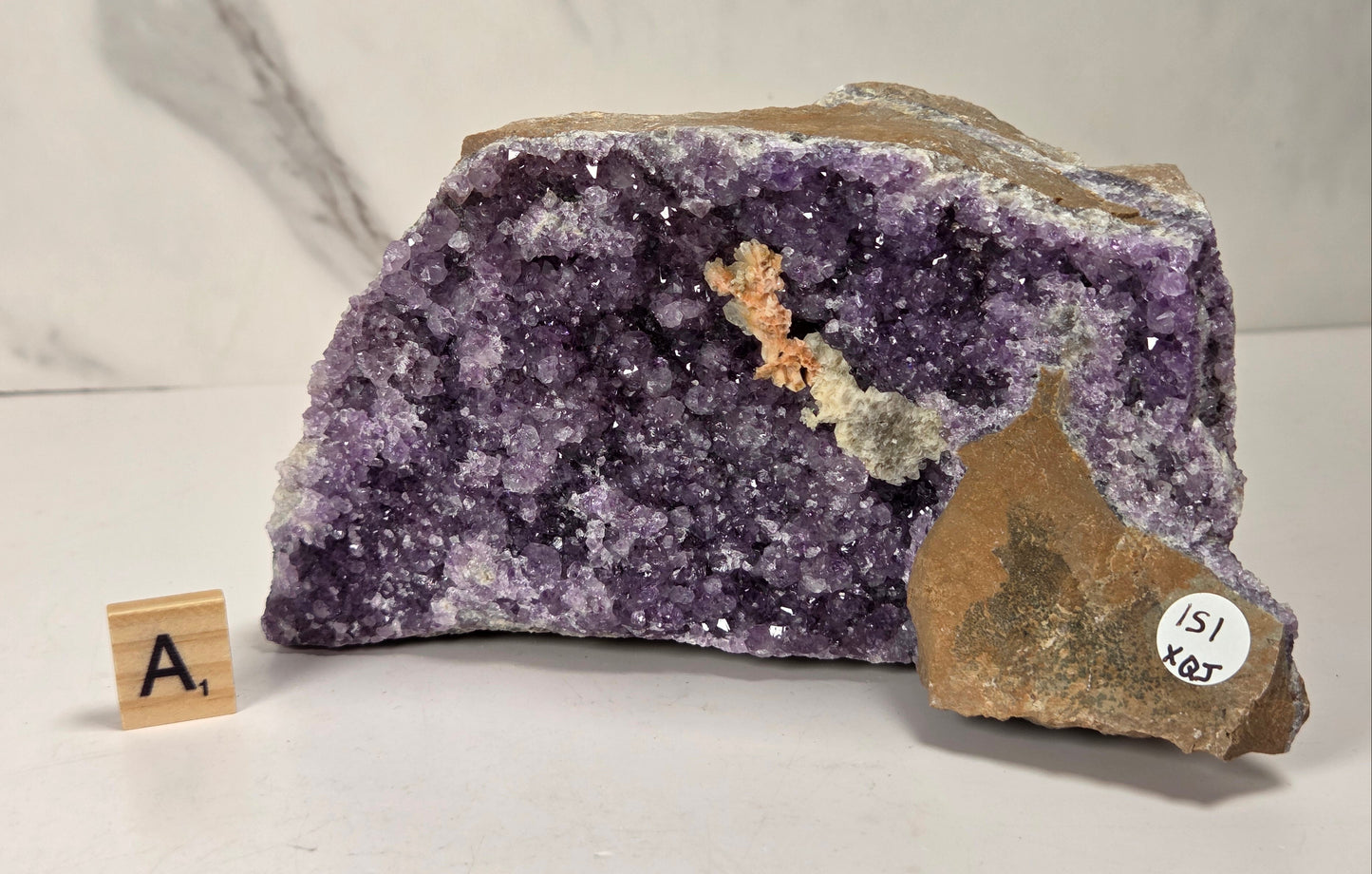 A raw amethyst geode specimen with a purple crystal cavity and brown matrix, displayed on a white surface with a small wooden letter tile and a small white inventory sticker.