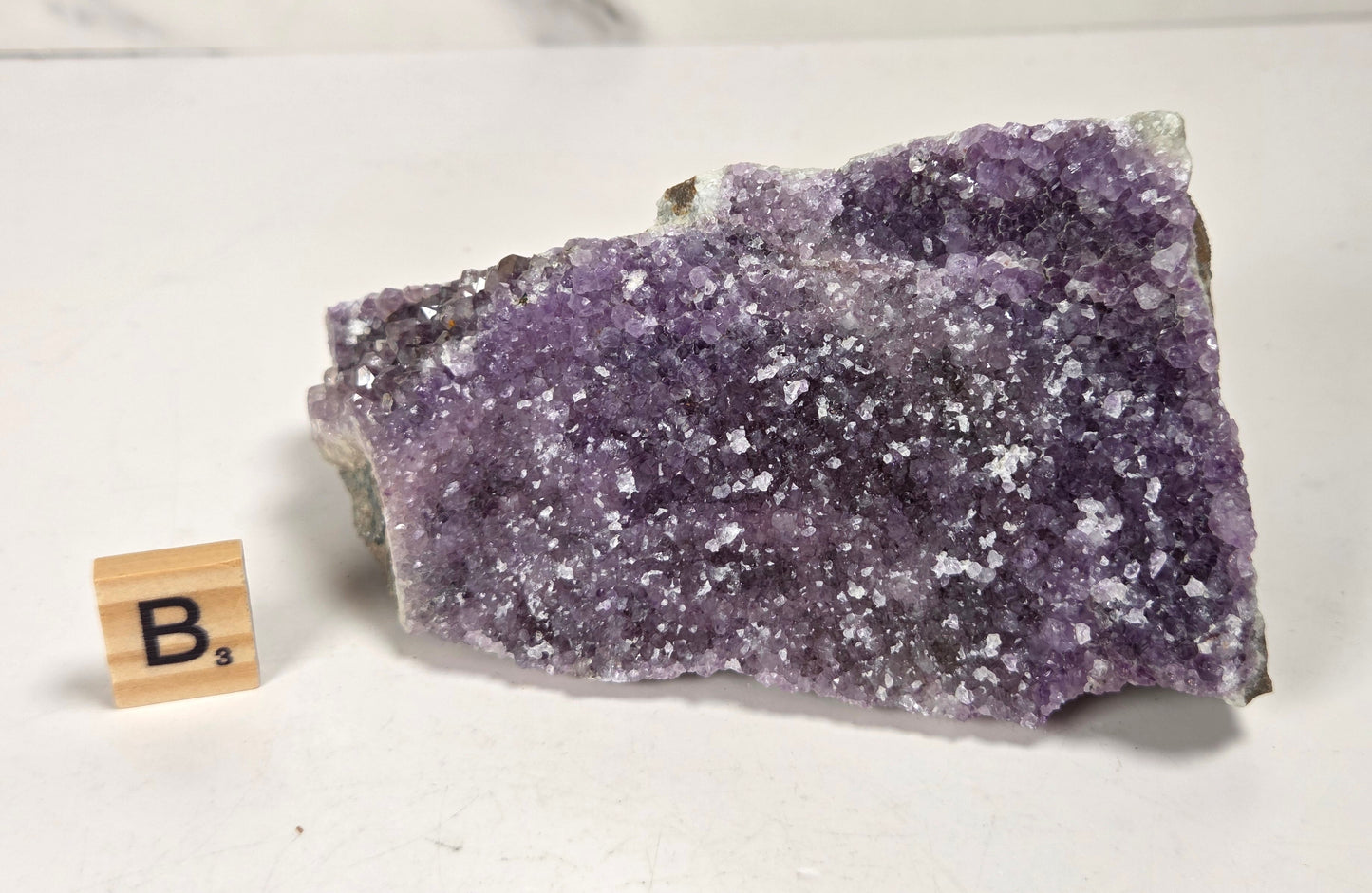 A raw amethyst crystal cluster with purple and white crystalline structure, displayed on a white surface next to a small wooden letter tile spelling 'B's'.