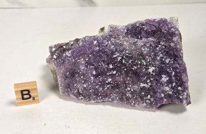 A raw amethyst crystal cluster with purple and white crystalline structure, displayed on a white surface next to a small wooden letter tile spelling 'B's'.