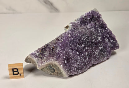 A raw amethyst crystal cluster with purple and white crystalline structure, displayed on a white surface next to a small wooden letter tile spelling 'B's'.