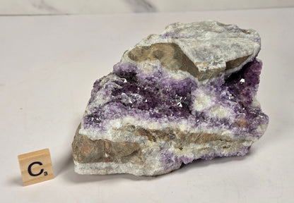 A raw amethyst geode specimen with purple crystal formations and a gray-brown matrix, displayed on a white surface with a small wooden 'C's' tile for scale.