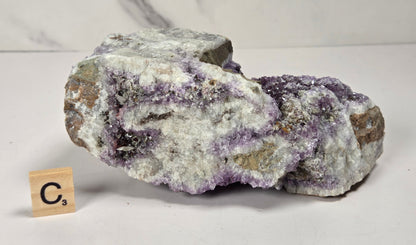 A raw mineral specimen with purple and white crystalline formations on a gray-brown matrix, displayed on a white surface with a small wooden 'C3' tile nearby.