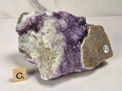 A raw amethyst geode specimen with purple crystal formations and a gray-brown matrix, displayed on a white surface with a small wooden 'C's' tile for scale.