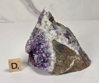 A raw amethyst geode specimen with purple crystal formations and a gray-brown matrix, displayed on a white surface with a small wooden 'D's' tile for scale.