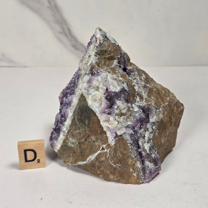 A raw mineral specimen with purple amethyst crystal points and white calcite crystals on a brown matrix, displayed next to a small wooden block with the letter 'D' on a white marble surface.