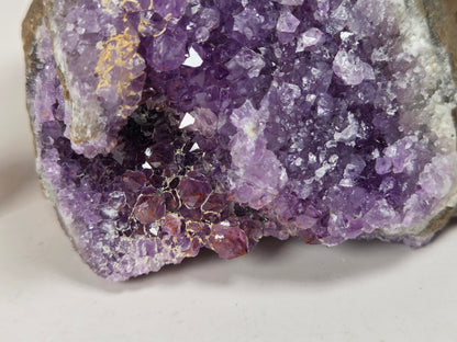 A raw amethyst geode specimen with purple crystal formations and a gray-brown matrix, displayed on a white surface 