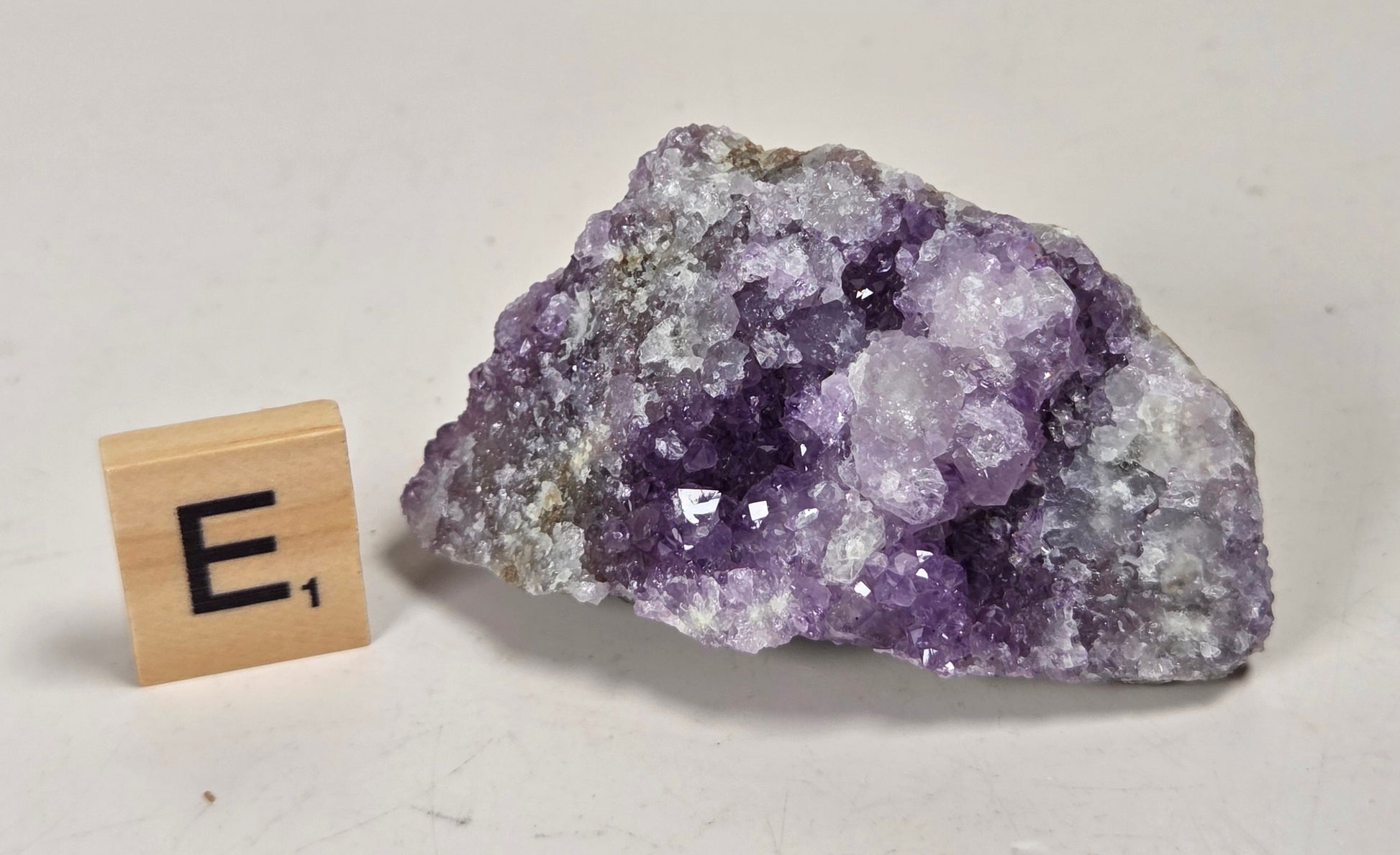 A raw amethyst geode specimen with purple crystal formations and a gray-brown matrix, displayed on a white surface with a small wooden 'E's' tile for scale.