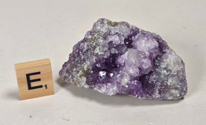 A raw amethyst geode specimen with purple crystal formations and a gray-brown matrix, displayed on a white surface with a small wooden 'E's' tile for scale.
