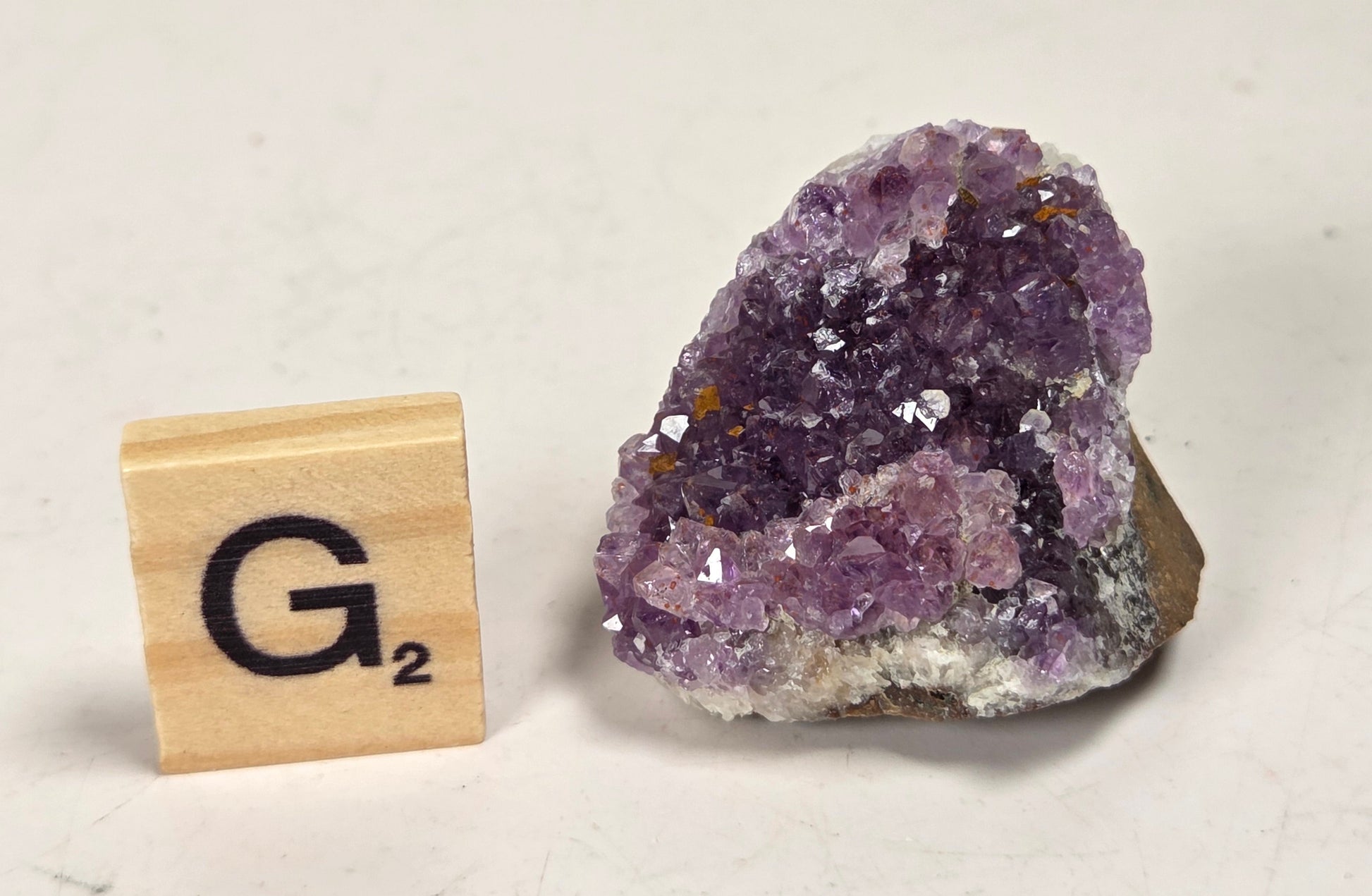 A small purple amethyst crystal cluster with a brown matrix, shown next to a wooden Scrabble tile marked G2 on a light off-white background.