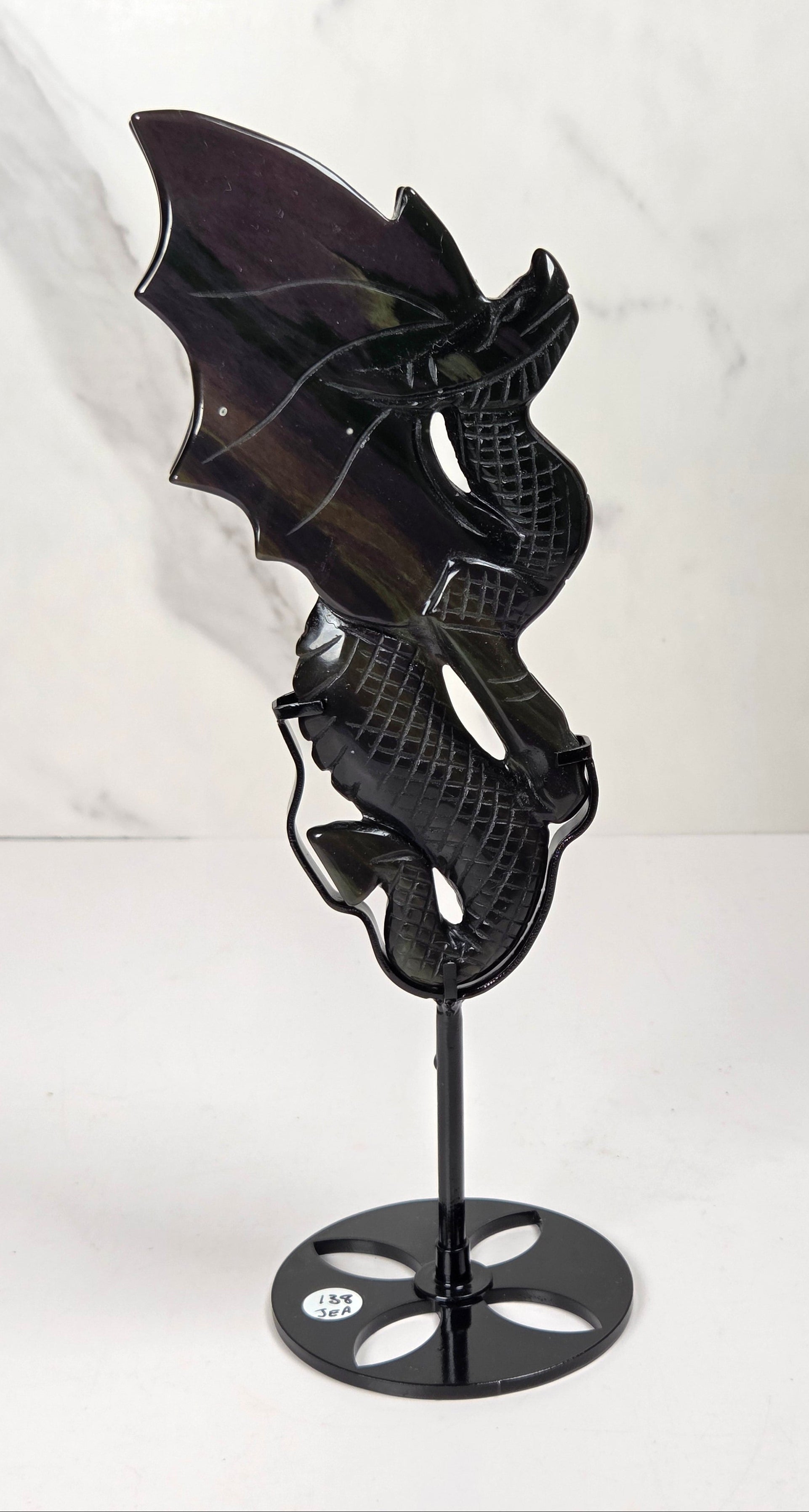 A black dragon figurine with spread wings mounted on a slender stand with a circular base, photographed against a light marble-patterned background.