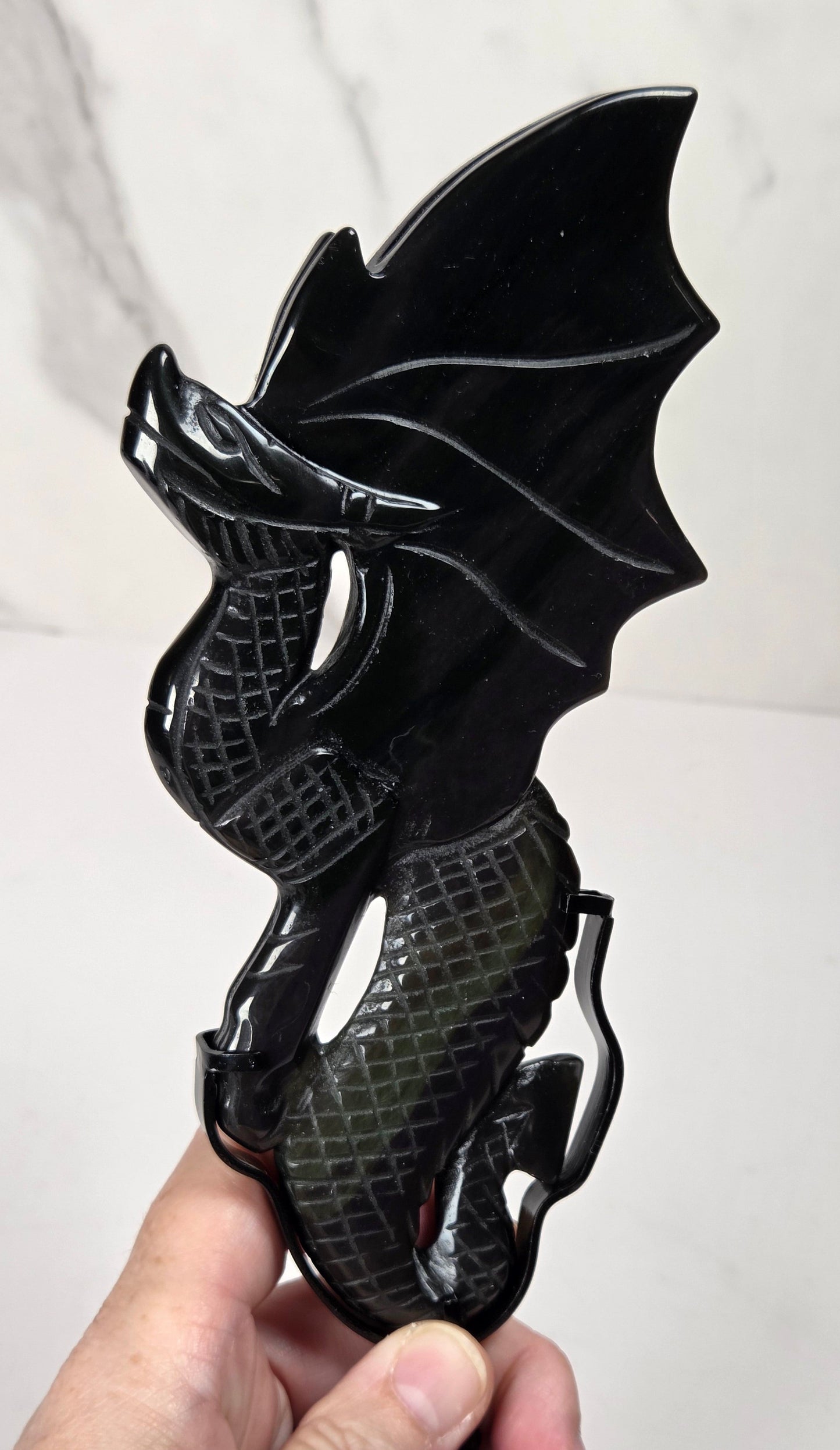 A hand holding a glossy black dragon figurine with a textured, scale-like body and bat-like wings, set against a light gray marble-patterned background.