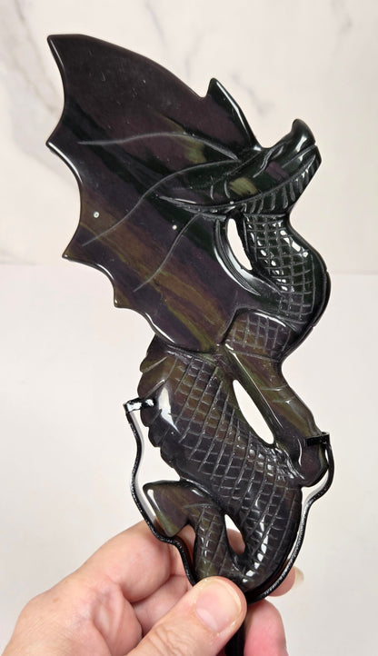 A hand holding a polished dark dragon figurine carved from what appears to be obsidian, with a mottled black, brown, and green sheen, set against a light gray marble-patterned background.