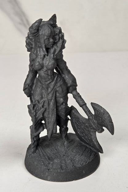 A dark gray unpainted fantasy miniature figurine of a female warrior holding a large double-headed axe, standing on a round base against a light gray and white marble-patterned background.