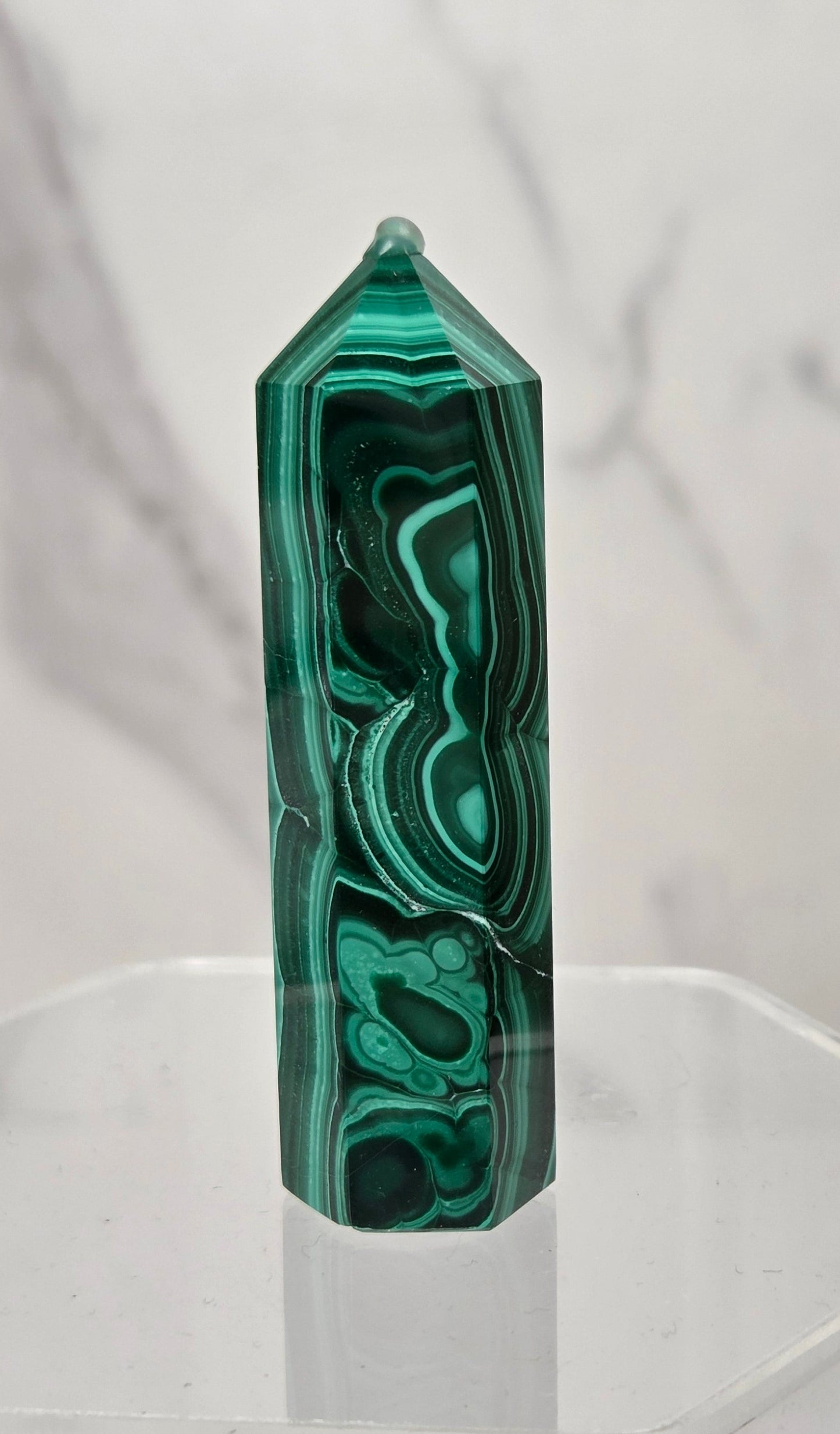 A polished green malachite crystal point tower displayed on a clear acrylic stand against a light gray marble-patterned background.