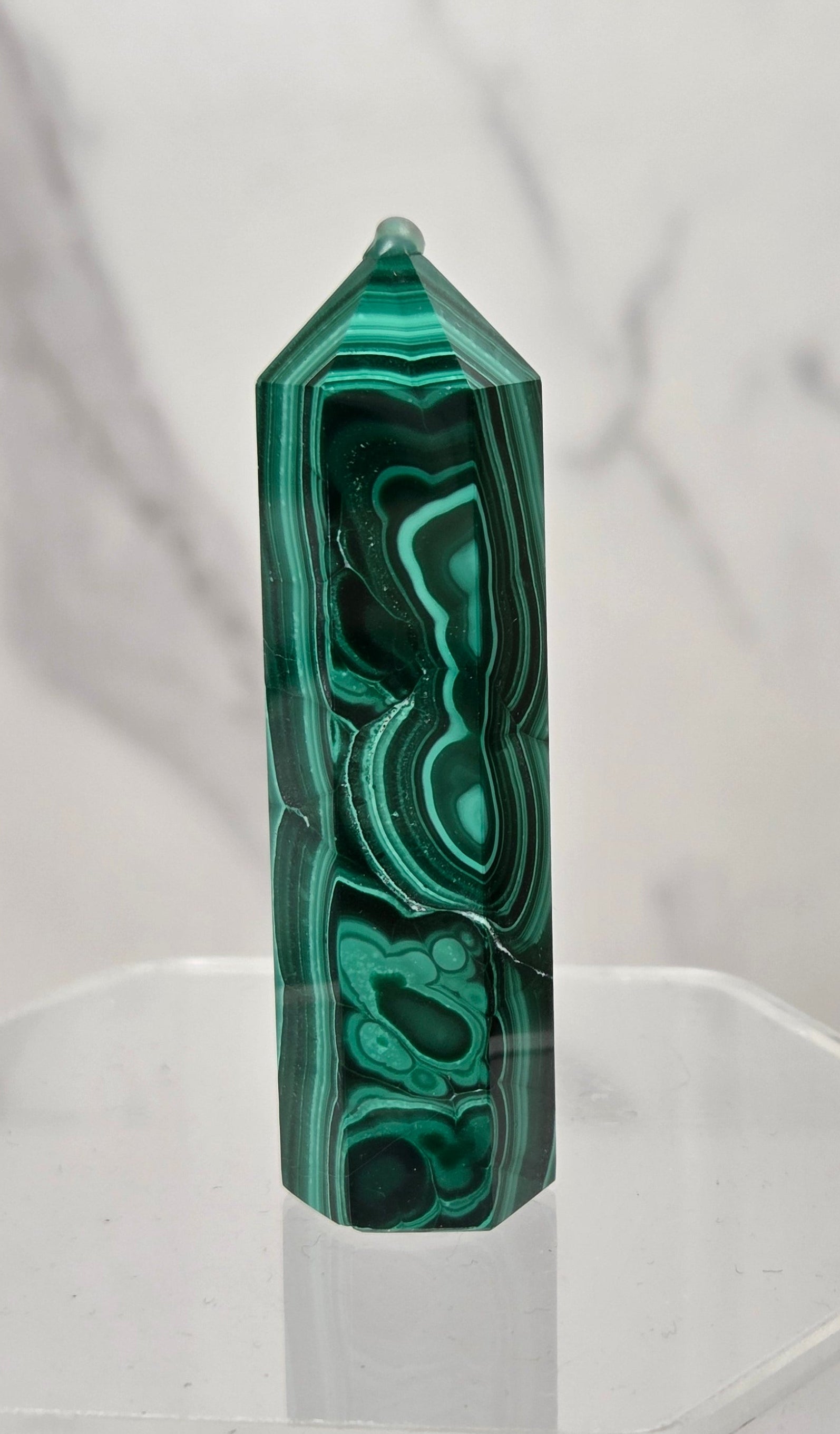 A polished green malachite crystal point tower displayed on a clear acrylic stand against a light gray marble-patterned background.