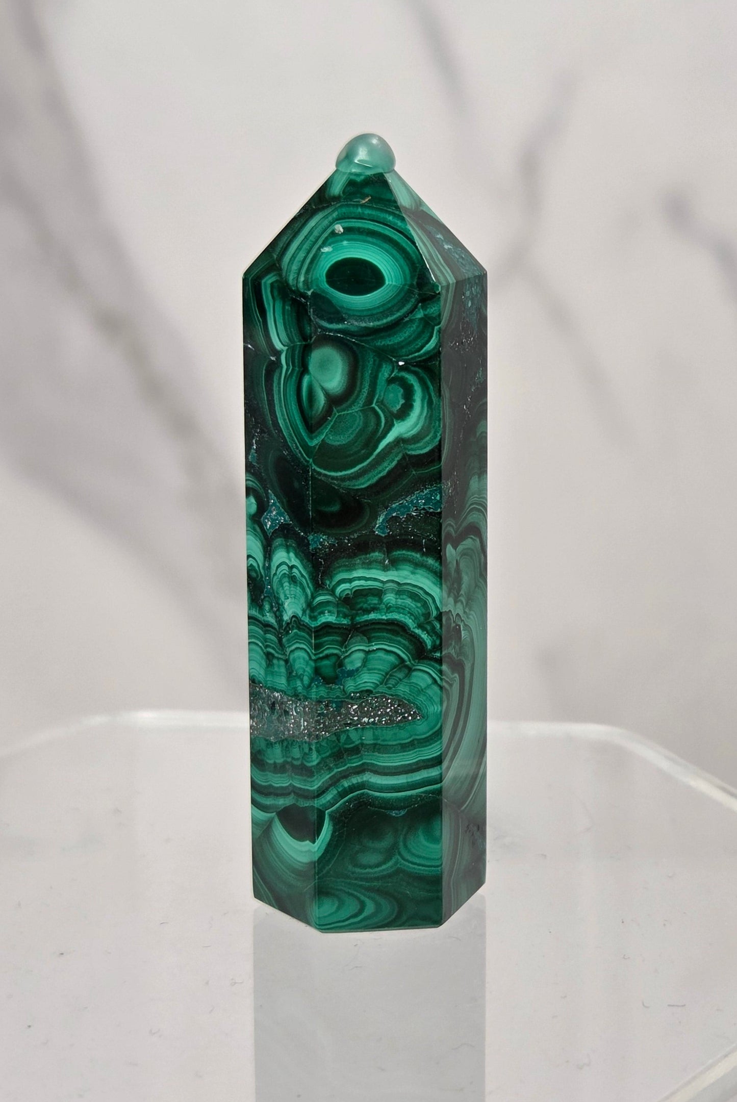A polished green malachite crystal point tower displayed on a clear acrylic stand against a light gray marble-patterned background.