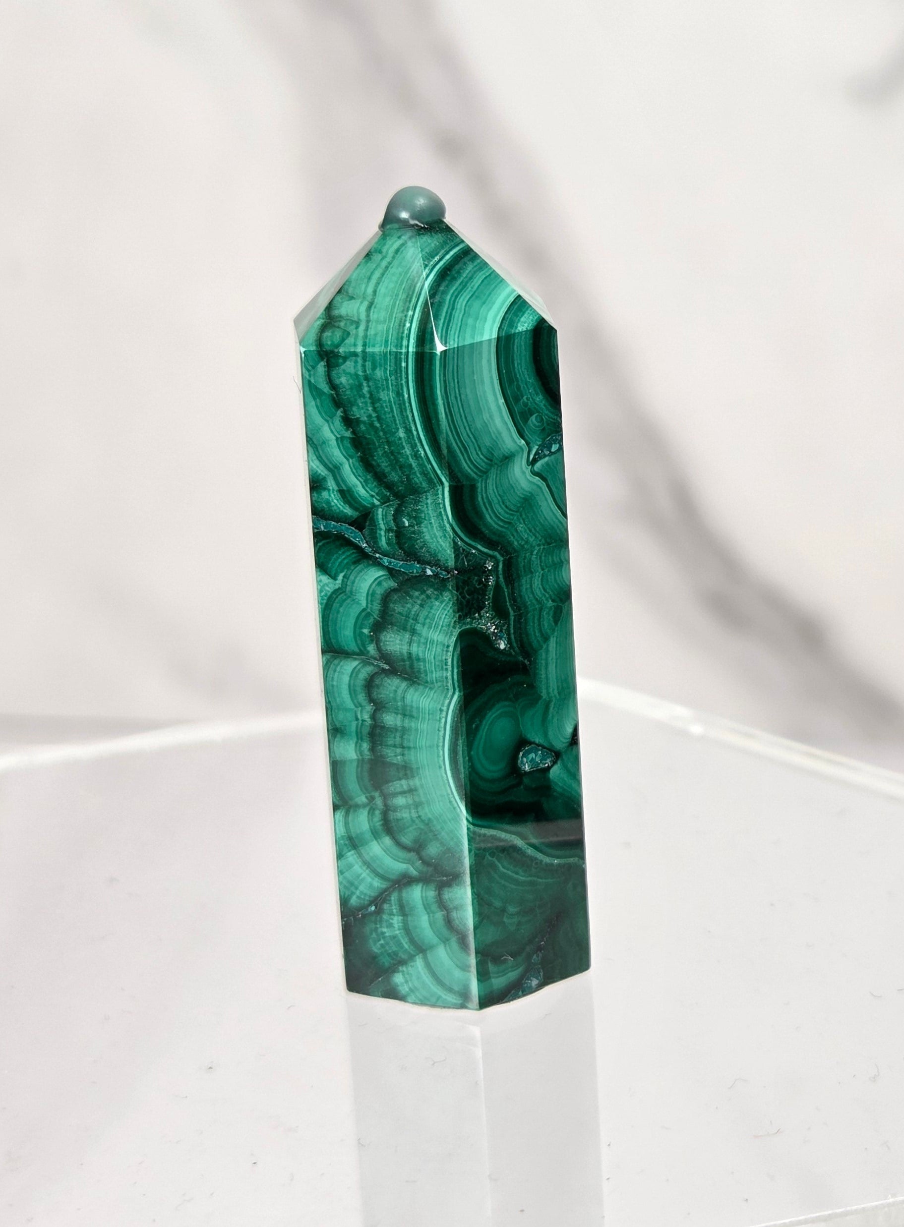 A polished green malachite crystal tower with natural banding patterns, displayed on a clear acrylic hexagonal stand against a white marble background.